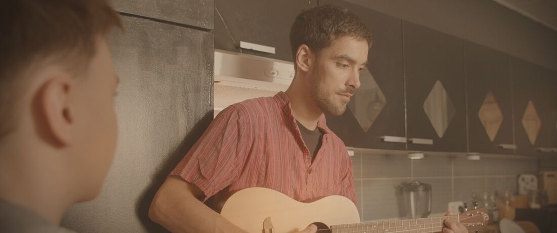 Un homme joue de la guitare dans une cuisine, observé par l’enfant au premier plan